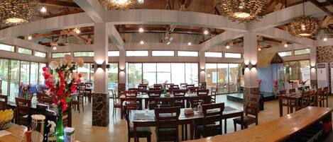 A spacious restaurant with multiple dining tables and chairs, large windows, and a central decorative floral arrangement.