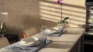 Un coin repas avec une longue table dressée pour deux personnes, agrémenté d’un vase contenant une seule orchidée, d’un verre d’eau et d’un bol posé sur un set de table à motifs.