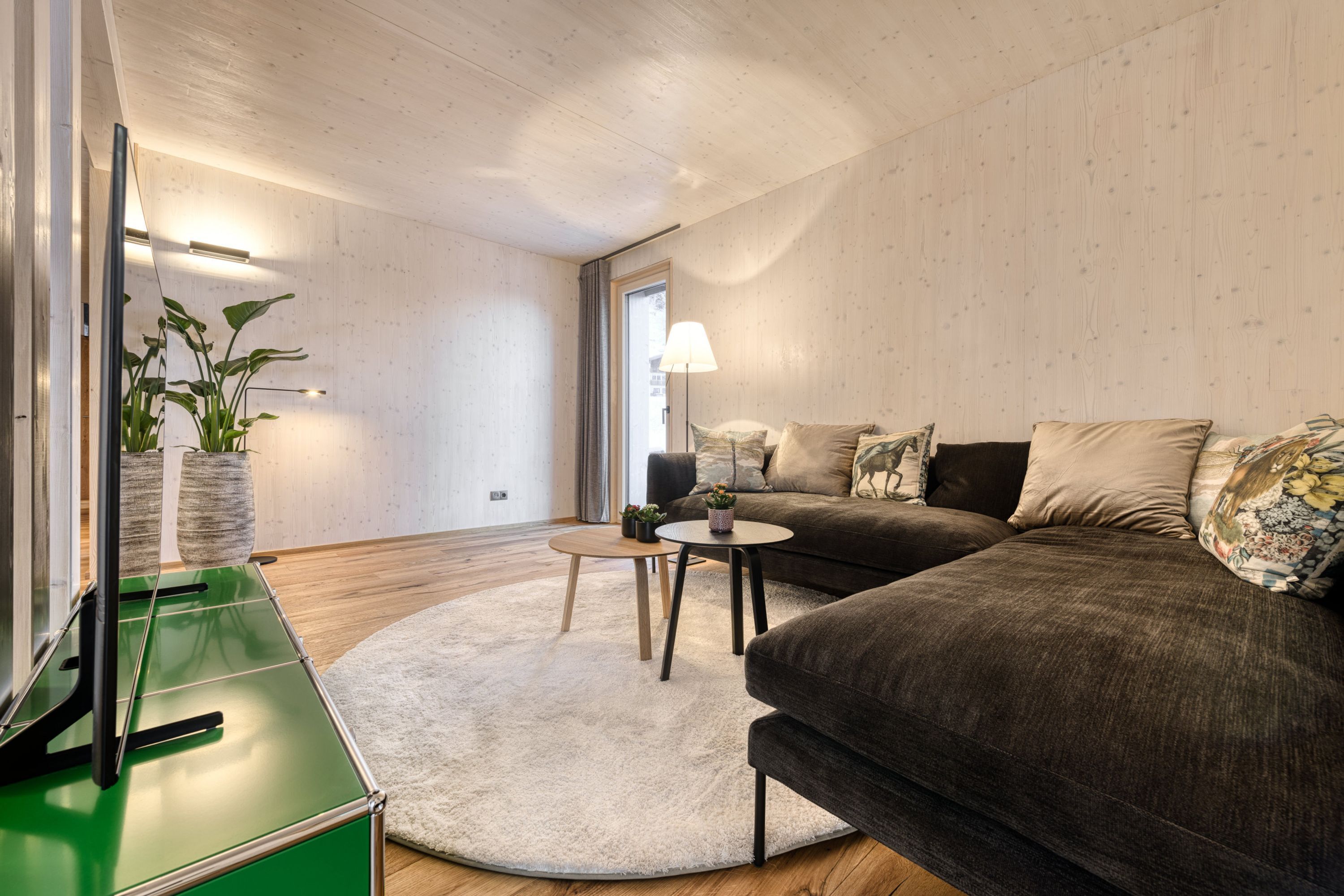 A modern living room with a green TV stand, a round rug, a dark sofa with patterned cushions, and a small table with a lamp.