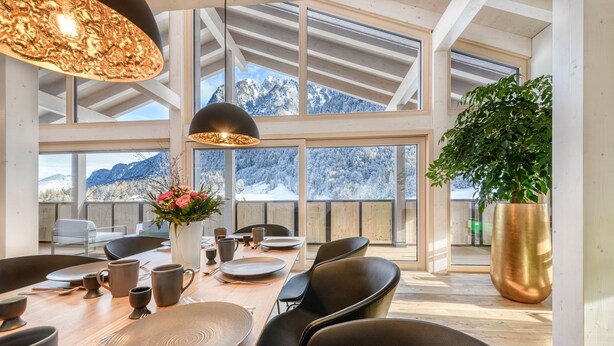 A dining area with a wooden table set for six, black chairs, and a large window offering a view of snow-covered mountains. - Luxus Penthouse Zentral (Brand)