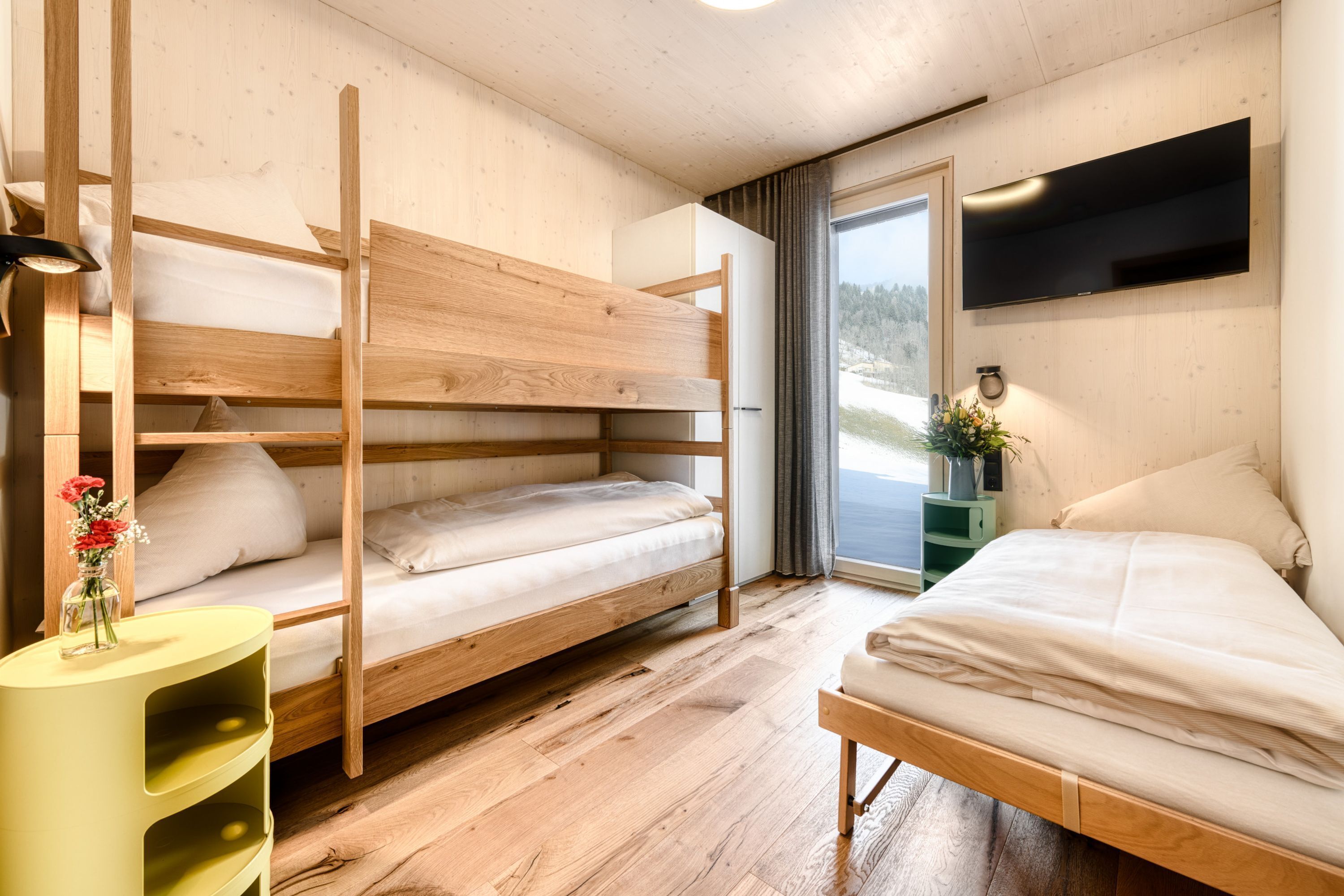 Ein Zimmer mit Etagenbetten, einem Flachbildfernseher, einem kleinen Tisch mit einer Vase und einem Fenster mit Blick ins Freie.