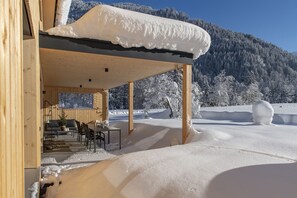 Una cabaña de madera con un porche cubierto, techo cubierto de nieve y vistas a un paisaje montañoso nevado.