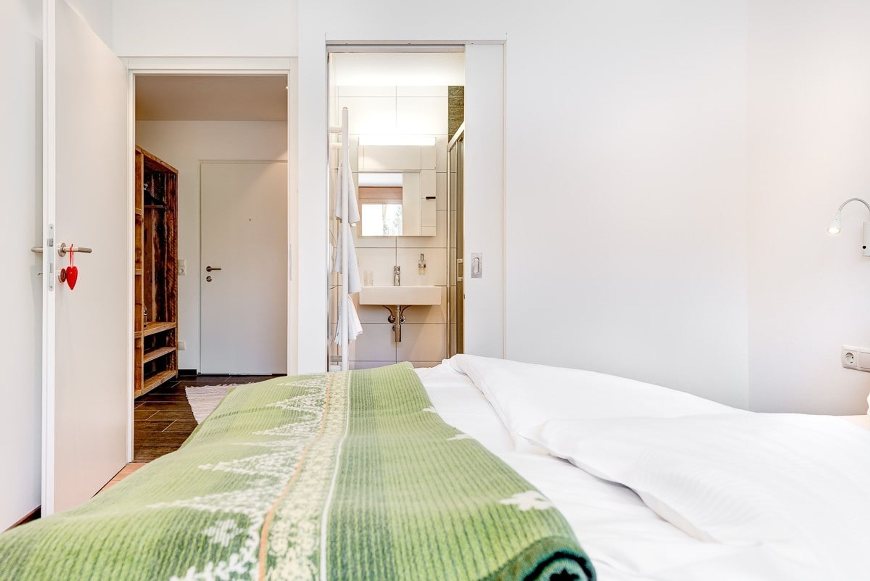 A hotel room with a bed, a green blanket, a wooden wardrobe, and a bathroom visible through an open door.
