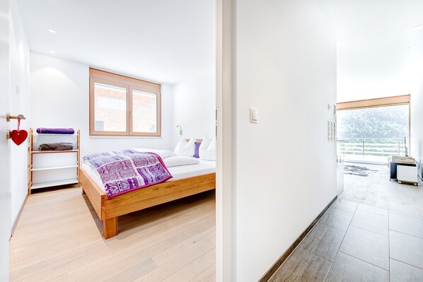 A bedroom with a wooden bed, a window, a shelf with towels, and a view of the outdoors. - Appartement Valbona Blick (Buerserberg)