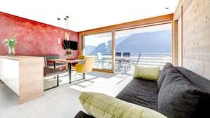 A modern living room with a sofa, a dining table with chairs, a small kitchen area, and a large window offering a view of mountains. - Appartement Panorama Blick (Buerserberg)
