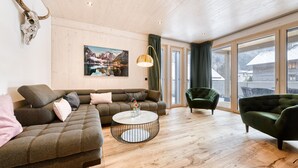 A modern living room with a sectional sofa, a round coffee table, and a green armchair. A large window offers a view of a snowy landscape. - Familienferienwohnung Zentral (Brand)