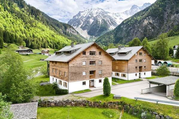 A multi-story building with a brick facade, a parking area, and a view of a mountainous landscape. - Gartenferienwohnung Murmele (Brand)