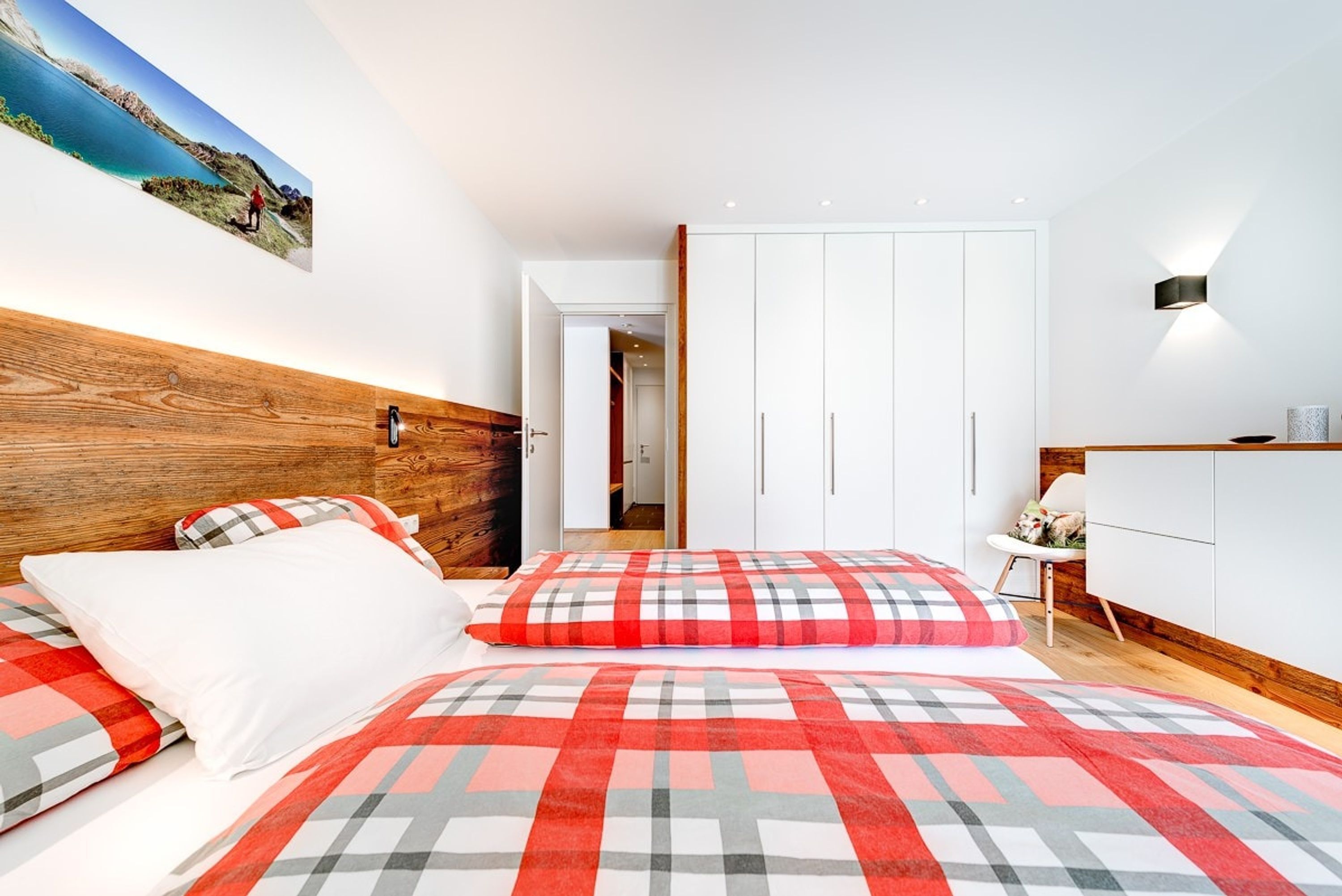 A bedroom with a wooden headboard, a bed with a red and grey checkered bedspread, white pillows, and a painting of a landscape on the wall.