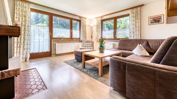 Un salon chaleureux avec un canapé, une table basse et une cheminée. De grandes fenêtres sont agrémentées de rideaux, une lampe et une plante ornent le rebord de la fenêtre.