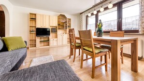 A modern living room with a sofa, dining table, chairs, wooden cabinets, a flat-screen TV, and a large window with curtains. - Appartement Golm - M Ser-apartments (Tschagguns)