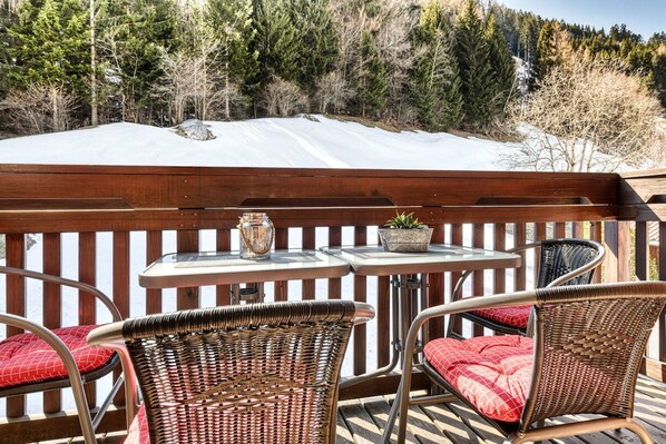 Una terraza de madera con dos sillas de mimbre y una mesita pequeña, con vistas a un paisaje nevado con árboles.