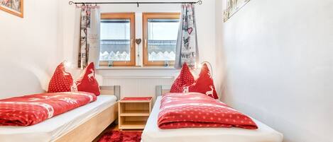 A room with two single beds, a small table, a red carpet, and a window with curtains.