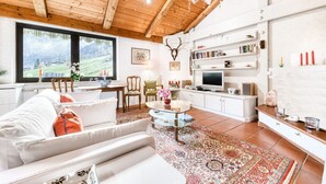 A cozy living room with a sofa, a coffee table, and a TV. There's a kitchen area with a stove, a sink, and a refrigerator. A large window offers a view of a green landscape. - Chalet Zentral Bach in Brand (Brand)