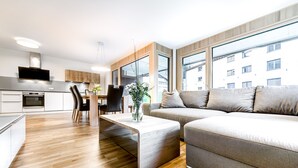 Un appartement moderne comprenant une cuisine, un coin repas et un salon. Il est doté d’un parquet, d’un canapé gris et de grandes fenêtres offrant une vue sur la ville.