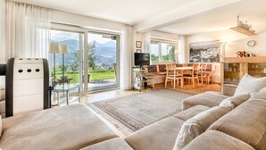 A spacious living area with a large sofa, a dining table, and a TV. There is a sliding glass door leading to a balcony with a mountain view. - Panoramavilla Bludenz (Bludenz)