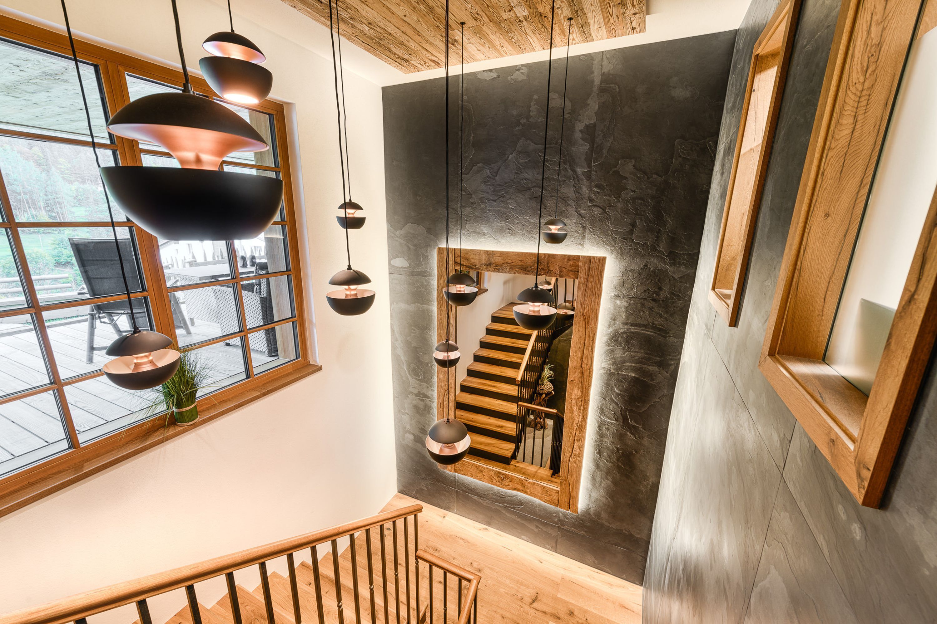 Ein modernes Interieur mit Treppe, Pendelleuchten und einem großen Fenster mit Blick auf eine Terrasse.