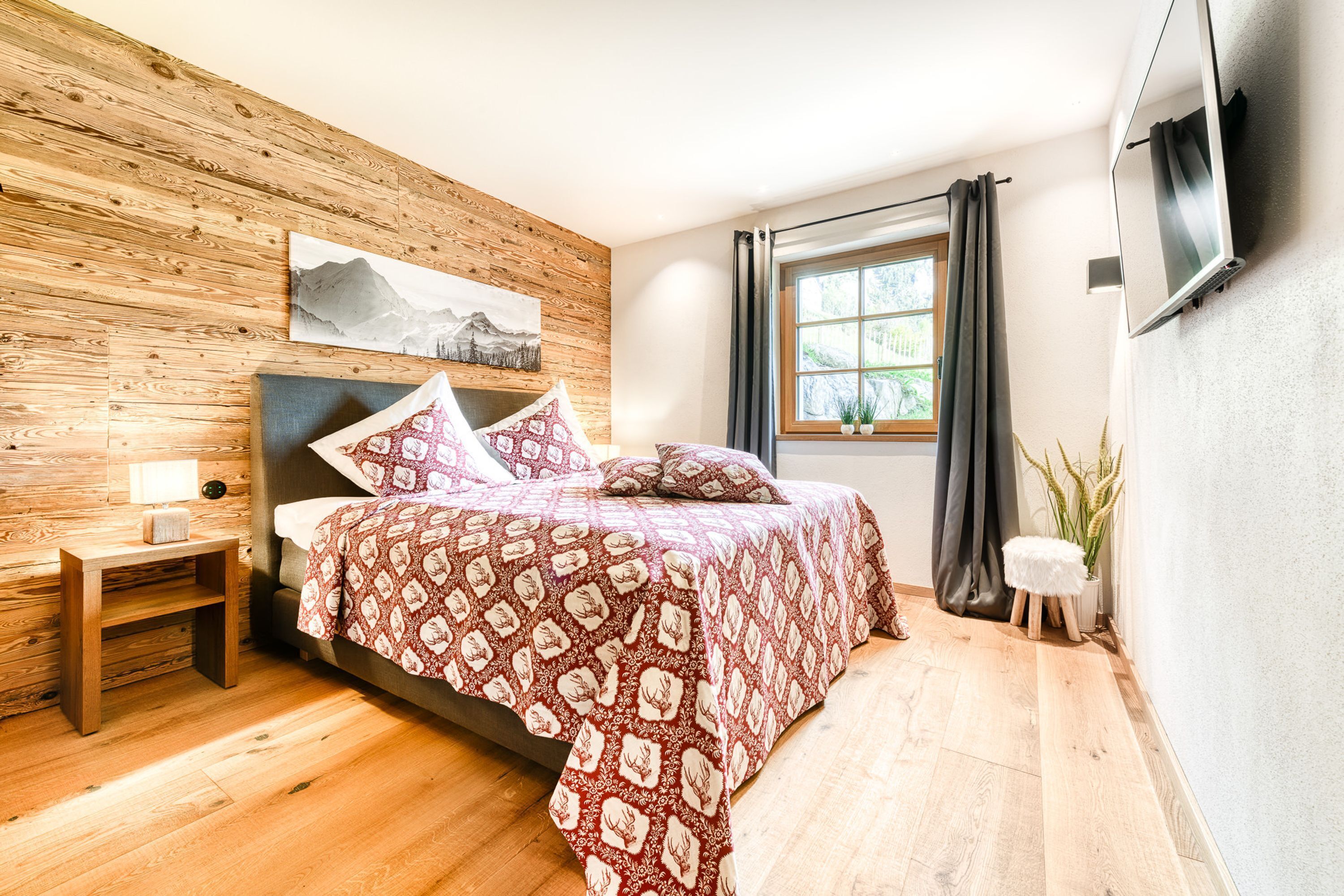 Ein Schlafzimmer mit einem Holz-Kopfteil, einem Bett mit gemusterten Betttextilien, einem Nachttisch, einem Fenster mit Vorhängen und einem angebrachten Fernseher.