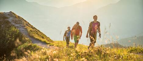 Un grupo de personas caminan por un sendero de montaña con vistas despejadas a montañas distantes y un sol brillante al fondo.