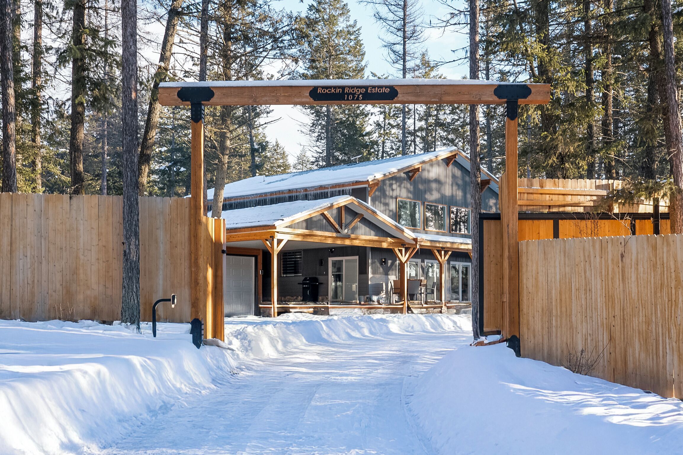 A wooden gate with a sign reading "Rockin Ridge Estate 1075" leading to a cabin with a metal roof and wooden beams.