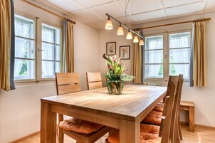 Un comedor con mesa de madera, sillas, un jarrón con flores y ventanas grandes con cortinas.