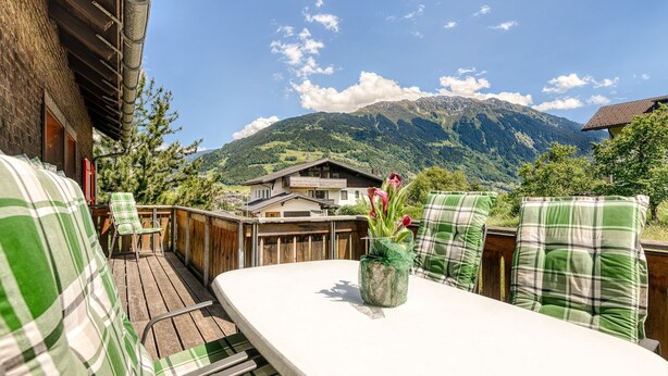 A wooden deck with a table and chairs, a view of mountains, and a house in the background. - Chalet Mariedl in Tschagguns (Tschagguns)