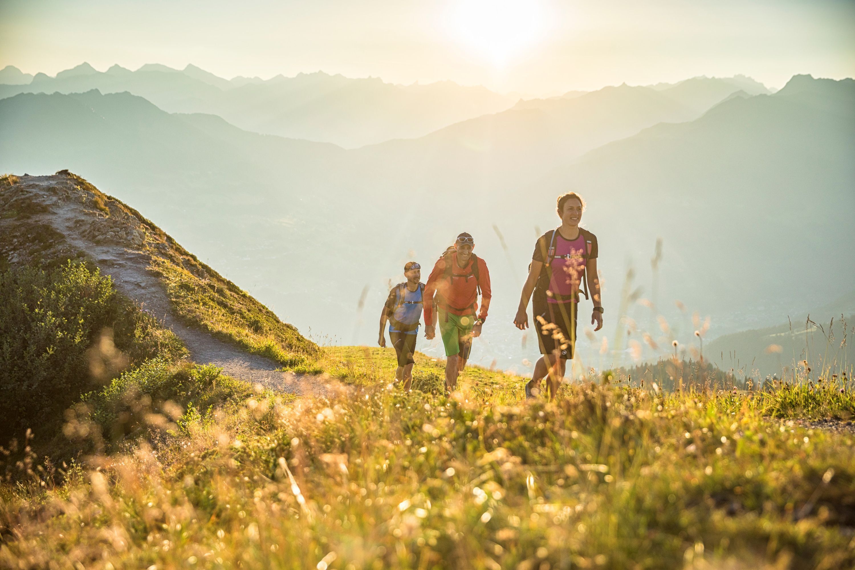 Un groupe de personnes fait de la randonnée sur un sentier de montagne, avec une vue dégagée sur les montagnes lointaines et un soleil éclatant en arrière-plan.