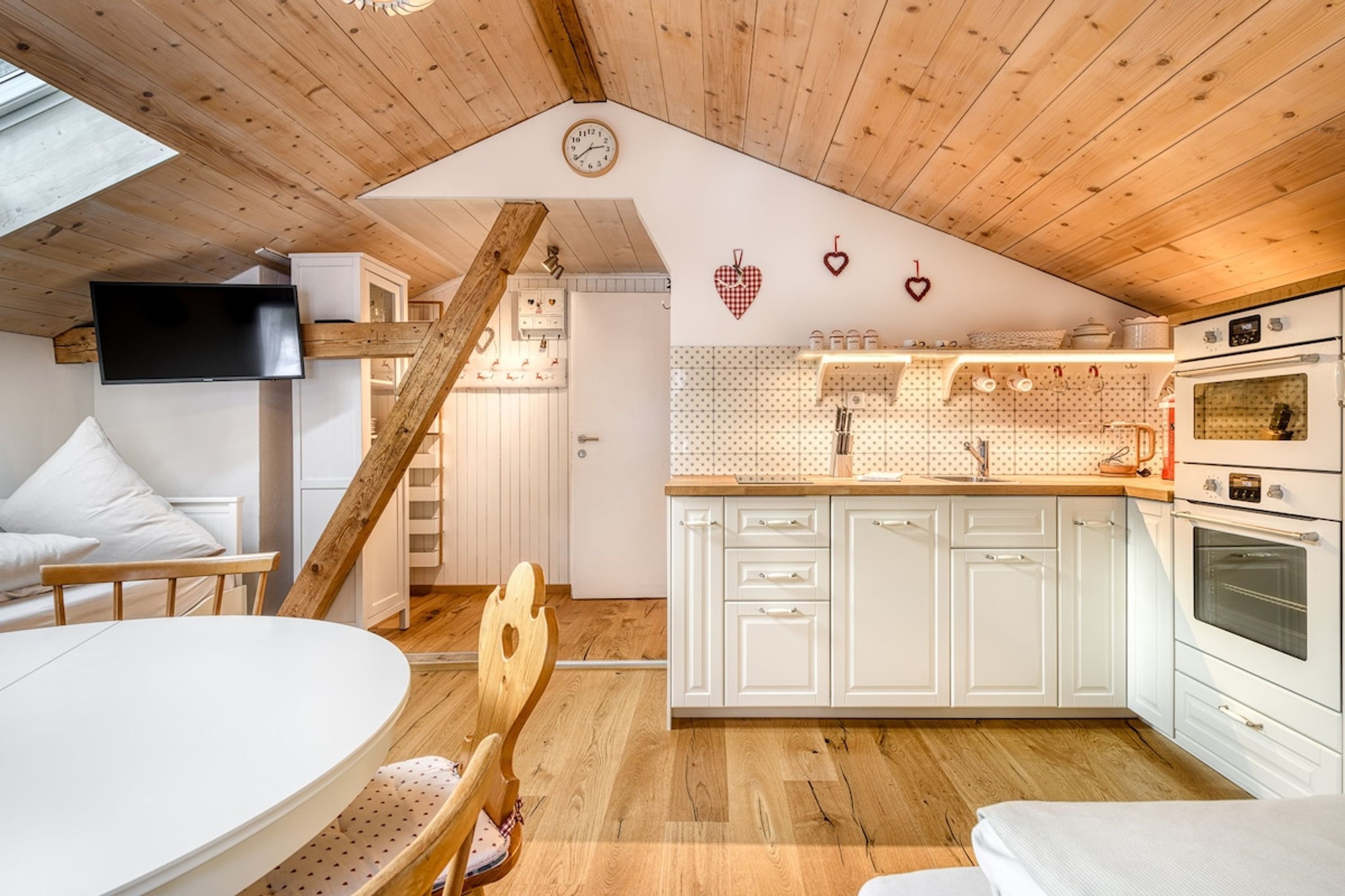 Une cuisine chaleureuse avec des poutres en bois, des meubles blancs, un escalier en bois et un téléviseur fixé au mur.
