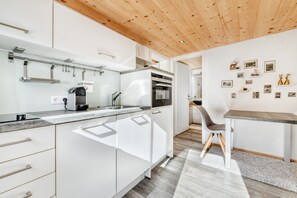 A modern kitchen with white cabinets, a wooden ceiling, and a dining area with a gray chair and table. - Ferienwohnung Zweisamkeit (Brand)