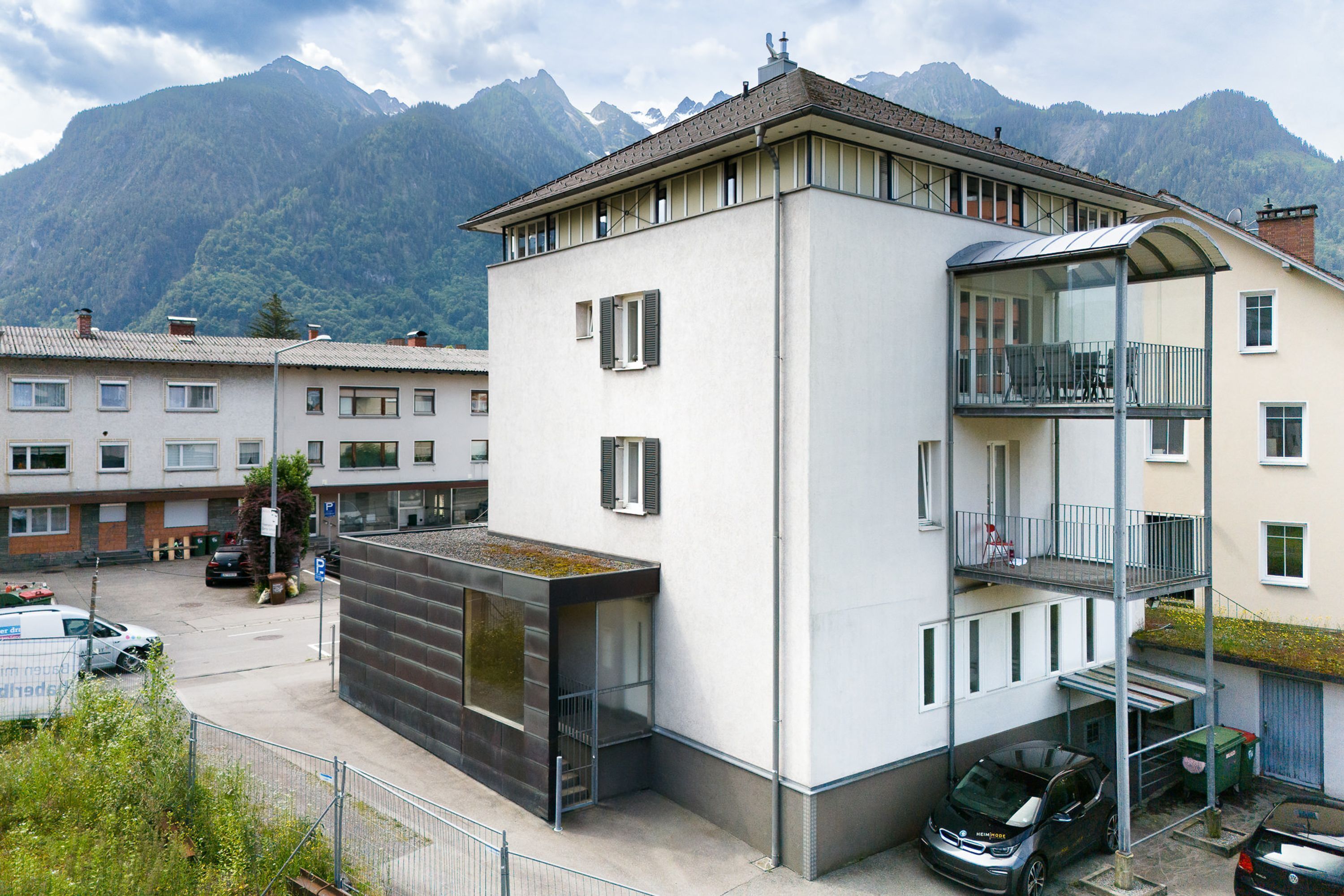 A modern building with a balcony, a carport, and a mountain backdrop.