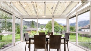 A covered patio with a table and chairs, large windows, and a view of a green landscape. - Landhaus Bergblick in Ludesch (Ludesch)