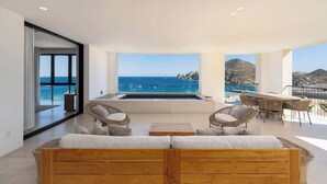 A modern living room with a large window overlooking the sea, a wooden sofa, wicker chairs, and a dining area with a table and chairs. - Luxury  Oceanfront Condo with Private Plunge Pool (Cabo San Lucas)