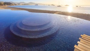 A clear view of an infinity pool with a wooden deck, overlooking a beach with boats and a distant shoreline. - Luxury  Oceanfront Condo with Private Plunge Pool (Cabo San Lucas)