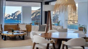 A modern living room with a large round table and white chairs, a hanging woven light fixture, and a view of the ocean and mountains through large windows. - Luxury  Oceanfront Condo with Private Plunge Pool (Cabo San Lucas)