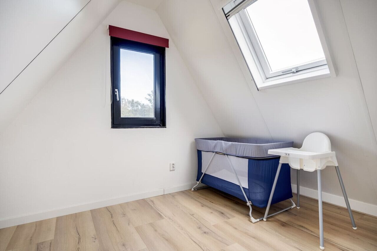 A room with a sloped ceiling, a window with a red frame, a blue and white mattress with a white chair, and a wooden floor.