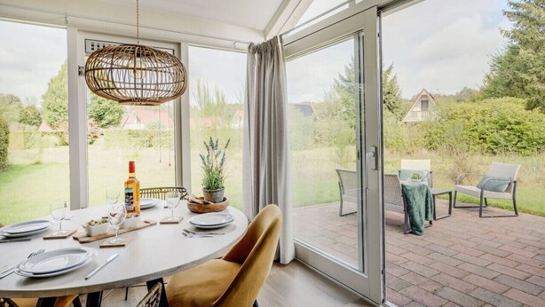 A dining area with a table set for four, a wicker pendant light, and a view of a garden and house through large glass doors. - Braamsluiper | Vakantiewoning met grote zonnige tuin (Zwiggelte)