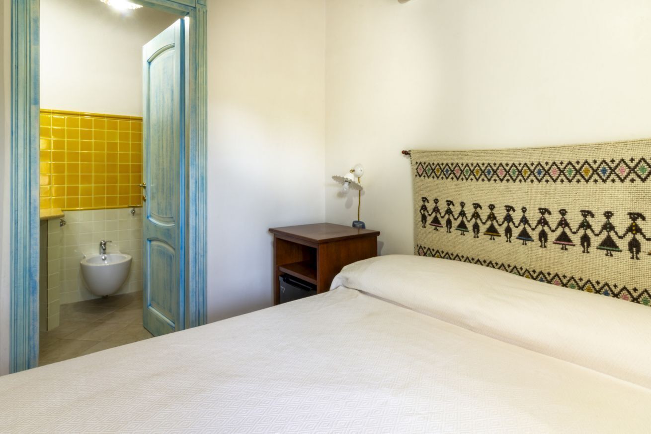 A hotel room with a bed, a nightstand, a small table, a wall-mounted shelf, a bathroom with a sink and a mirror, and a door leading to the bathroom.