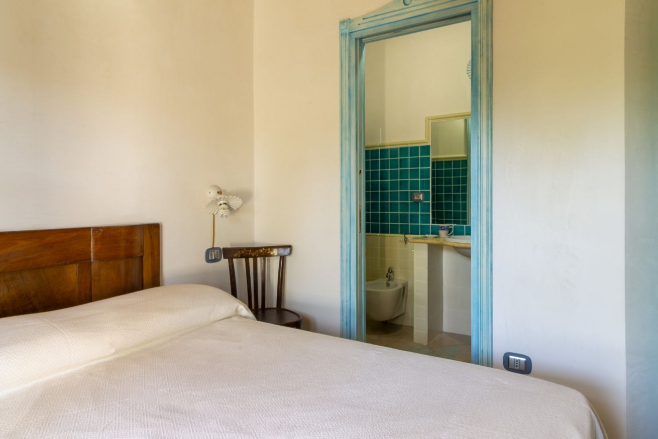 Een slaapkamer met een houten hoofdbord, nachtkastje en stoel. Een badkamer met een wastafel en spiegel is door een open deur zichtbaar.