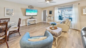 A living room with a television, a ceiling fan, a dining area with chairs, a sofa, and a window with a view of a building. - PCB Beach Retreat with Pool and Private Balcony (Panama City Beach)