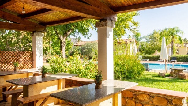 Un espace extérieur aménagé en terrasse, avec des colonnes en pierre, des bancs en bois et une piscine en arrière-plan.