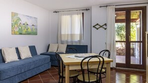 Une pièce avec un canapé bleu, une table en bois entourée de chaises, un tableau accroché au mur et une porte donnant sur l’extérieur.