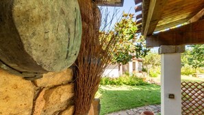 A stone wall with a broom leaning against it, a rustic wooden pergola, and a garden with a stone pathway and a large clay pot.