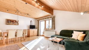 A cozy living room with a green sofa, wooden dining table, and chairs. There is a television and a clock on the wall. - Haus Alvier - 2-zimmer Fewo Zimba mit Balkon (Brand)