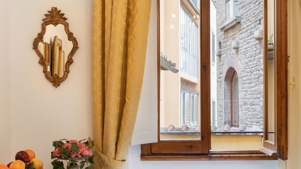 Una stanza con un tavolo in legno, uno specchio, una finestra che si affaccia su un edificio in pietra e un vaso con fiori.