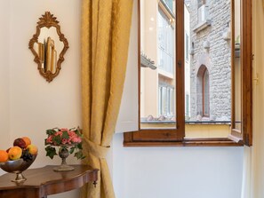 Um quarto com uma mesa de madeira, um espelho, uma janela com vista para um edifício de pedra e um vaso com flores.