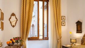 Camera d'albergo con un letto, un tavolo con una ciotola di frutta, una lampada e vista su un edificio in pietra dalla finestra.