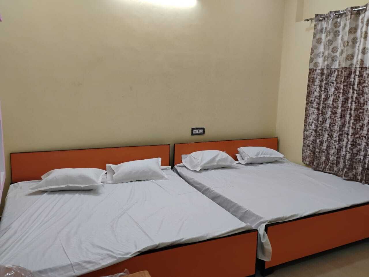 Cama doble con ropa blanca y cabecera naranja, un enchufe en la pared y una cortina estampada en la ventana.