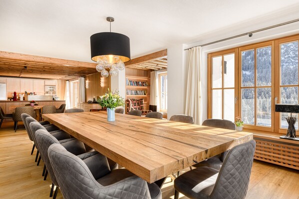 A spacious dining area with a long wooden table surrounded by chairs, a black pendant light, and a bookshelf filled with books.