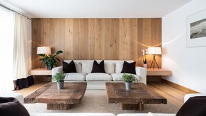 A modern living room with a wooden panel wall, a white sofa with dark cushions, two wooden coffee tables with plants, and a floor lamp. - Chalet Lilly-family Friends mit Ski In-ski out (Lech am Arlberg)