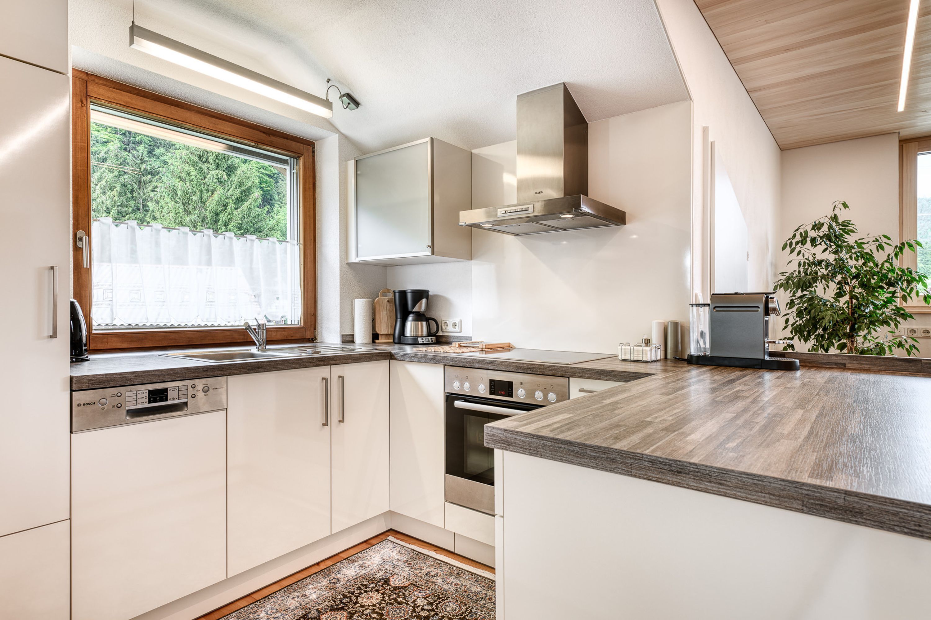 A modern kitchen with built-in appliances, a window with a view of greenery, and a wooden ceiling.