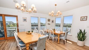 A dining area with a wooden table and white chairs, large windows with a view, and a ceiling light fixture.